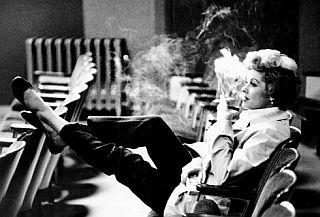 Lucille Ball with cigarette observing a rehearsal or reading of some kind, likely in a Desilu studio or sound stage. 