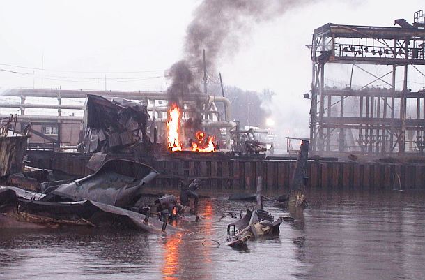 Smoldering remains and aftermath of the Bouchard Barge 125 explosion at ExxonMobil’s Staten Island depot. Half-sunken remains of the barge visible in the foreground, as large portions of it were blown out into the depot.