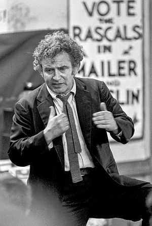 Norman Mailer in 1969, when he ran for Mayor of New York, in front of campaign poster and slogan for he and running mate Jimmy Breslin, “Vote the Rascals In.”