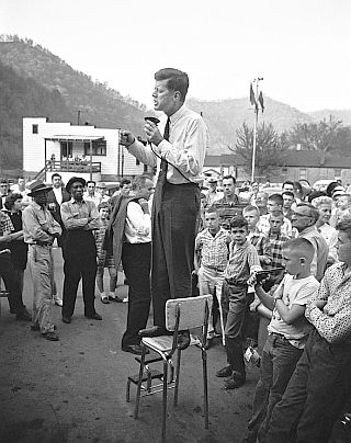 April 1960: JFK perched precariously on high chair as he addresses a gathering in Logan County, West Virginia during that state’s primary election. Photo, Hank Walker.