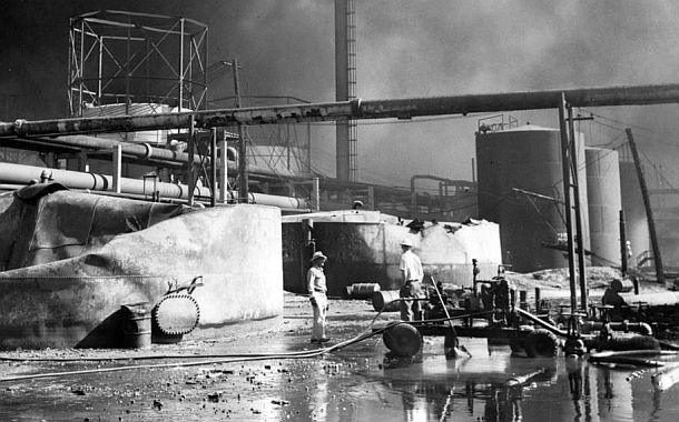 Two men at center survey the damage in a portion of the burnt-out Standard Oil refinery, then still smoldering, with the remains of two crumpled and distorted giant storage tanks to the left. Whiting oil refinery, Aug 1955, Chicago Tribune photo.