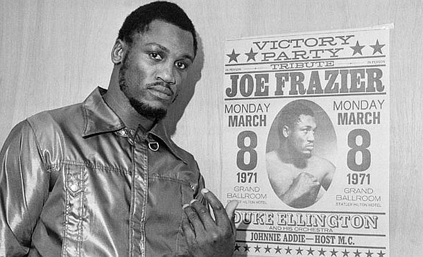 Heavyweight boxing champ, Joe Frazier, in 1971. By this time he had a 26-0 record, with 23 KOs; shown here posing by “victory party” poster after final public workout in Philadelphia, March 6, 1971.  AP photo/Bill Ingraham.