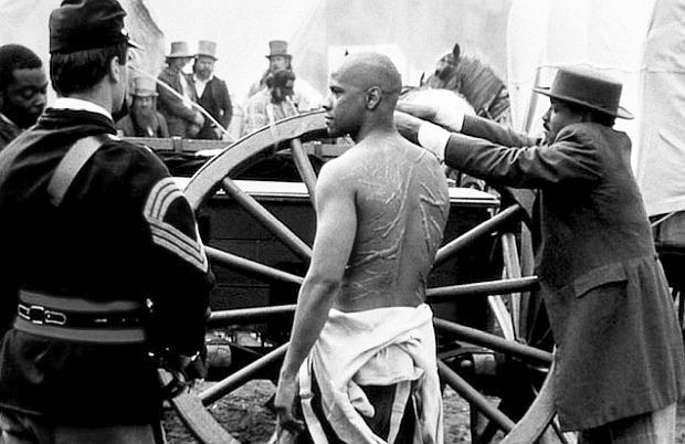 Pvt. Trip being tied to a wagon wheel prior to being flogged for alleged desertion, revealing  scars from his slave days and earlier whippings. Colonel Shaw later learns the real reason Trip left camp.